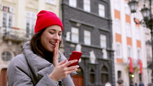 Portrait of Beautiful Woman in red hat Wearing warm closes Walking in the Town with Smartphone. Winter mood. Modern Mobile phone in her hands. Having online Conversation. Social Media Apps.