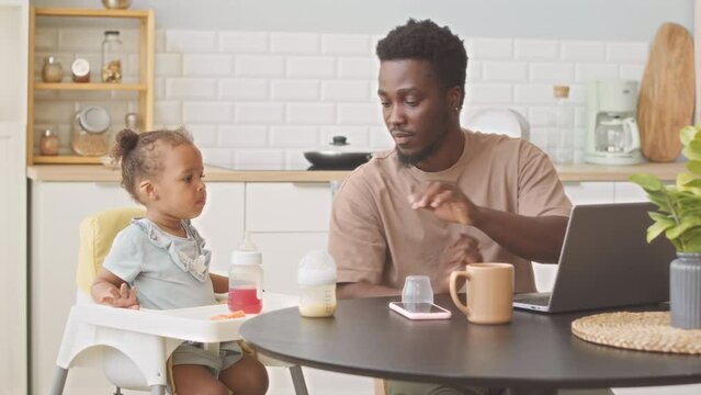African American man working on laptop at kitchen table while his cute toddler daughter sitting in high chair nearby having breakfast
