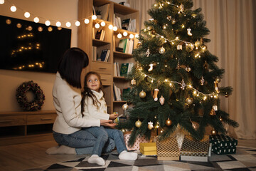 Happy small preschool caucasian child girl decorating Christmas tree with happy young mother, putting toys on branches, enjoying preparing for New Year celebration at home, miracle time concept.