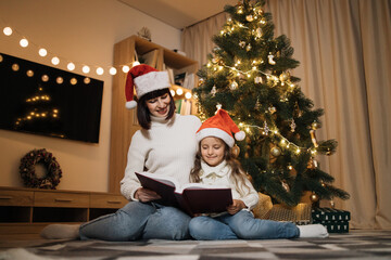 Happy family, attractive young mother and preschool little daughter in santa hat reading fairy tale book girl sitting near tree at evening at home. Merry Christmas concept.