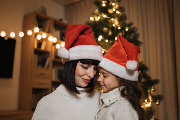 Fototapeta na wymiar Portrait of happy mother and little cute daughter in santa hat and warm knitted white sweaters sitting near decorating tree in living room. Loving family indoors. Merry Christmas and Happy Holidays.