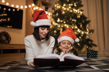 Fototapeta na wymiar Pretty young mother in santa hat reading magical fairy tale book to her daughter lying on floor near Christmas tree, merry Christmas.