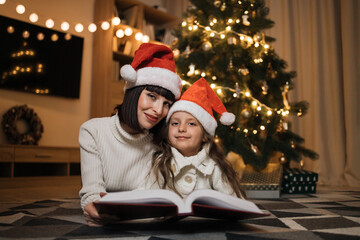 Close up view of lovely family of two, beautiful young mother and her cute little daughter in warm knitted white sweaters reading fairy tale book while lying in front of decorated Christmas tree.
