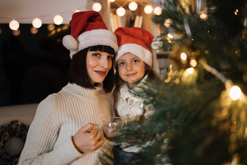 Fototapeta na wymiar Close up portrait of lovely family, beautiful young mother and her cute little daughter in warm knitted white sweaters drinking hot cocoa or chocolate from cups in front of decorated Christmas tree.