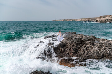 A woman stands on a rock in the sea during a storm. Dressed in a white long dress, the waves break on the rocks and white spray rises.