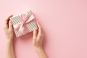 Christmas Eve concept. First person top view photo of young girl's hands holding stylish giftbox with bow on isolated pastel pink background with empty space