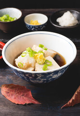 Fresh hot tofu in flavorful dashi and soy sauce-based brot served with  topping daikon, green onion,  chive ginger, autumn leaves over wooden table. Autumn and winter hot Japanese recipe