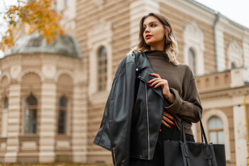 Fototapeta na wymiar Beautiful fashionable young girl in a stylish black leather jacket with a green sweater with a bag walks in the autumn city with a vintage building