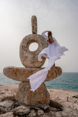 A woman stands on a stone sculpture made of large stones. She is dressed in a white long dress,...