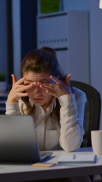 Vertical video: Stressed businesswoman suffering from headache at work doing overtime late night. Exhausted tensed worker coping with migraine, emotional stress concept feeling pain sitting at office