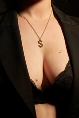 Cropped shot of a lady wearing a black lace bra, a black jacket and a metal golden necklace made out of a chain with a dangling pendant with a letter "S". The photo is made on the black background.