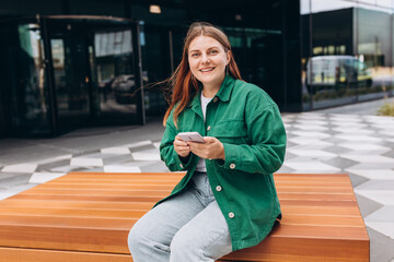 Young happy woman smiling and using smart phone at the city. Beautiful woman siting on bench on city street and searching information on the internet with smartphone. Business concept