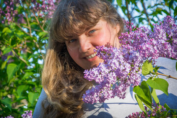 On a sunny day in the spring, a happy girl is standing on the street near the lilac. Close-up.