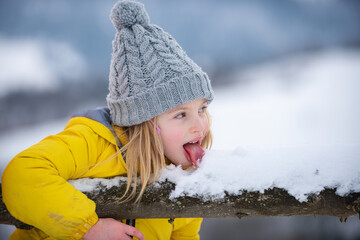 Winter girl eating snow outdoor. Funny christmas kids lick snow with tongue. Kids cold and flu concept. Children wearing warm winter clothing in snow weather. - obrazy, fototapety, plakaty