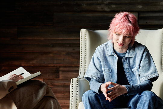 Teenage girl with pink hair sitting on chair at psychotherapy and talking to psychotherapist