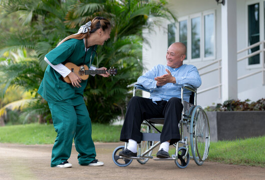 Asian female caregiver in green is singing and playing an ukulele with an elderly man on a wheelchair in a nursing home, a nursing care.
