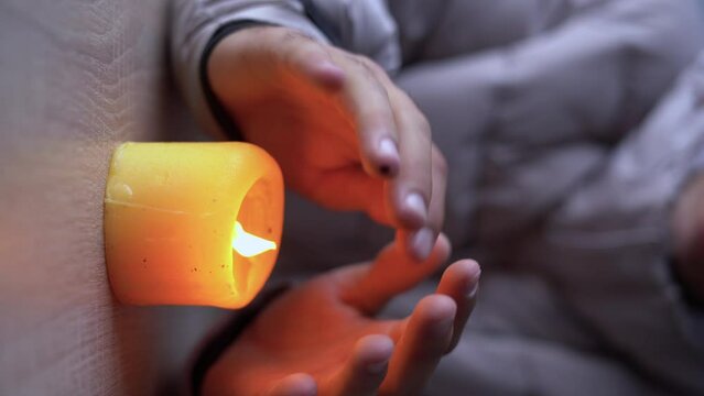 Close up hands of man dressed in a warm winter jacket sits at home at a table and warms his hands from a burning candle. no heating in winter at home