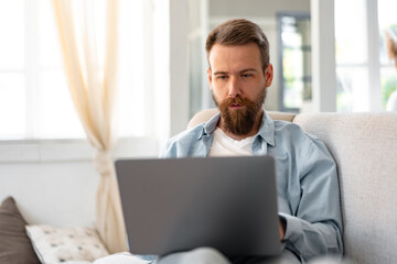 Young bearded man sitting on sofa at home using laptop computer