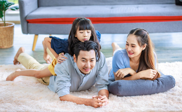 Millennial Asian lovely happy family father mother laying down on carpet floor while young daughter girl sitting piggy back on dad smiling laughing playing together in living room at home in weekend
