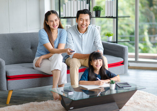 Millennial Asian happy family father and mother sitting on cozy sofa couch holding remote watching television while little young girl daughter sitting on floor doing homework in living room at home - Powered by Adobe