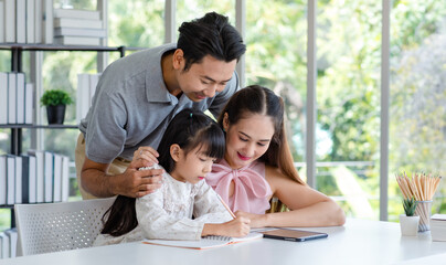 Millennial Asian happy family father and mother smiling helping supporting teaching little girl daughter studying learning writing doing school homework via tablet computer in living room at home