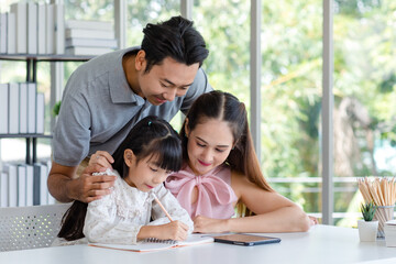 Millennial Asian happy family father and mother smiling helping supporting teaching little girl daughter studying learning writing doing school homework via tablet computer in living room at home