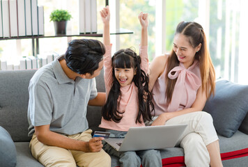 Asian happy excited little girl daughter sitting on cozy sofa couch smiling yelling screaming while father and mother using credit card making payment shopping gift present online via laptop computer