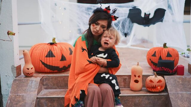Multiethnic family - mother and little son celebrate Halloween at home at background of decorated with pumpkin lanern house. Sweet unhappy boy in skeleton costume crying. Sad lonely holiday.