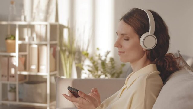 Happy young Caucasian woman enjoying music in headphones while chilling on sofa in spacious sunlit living room