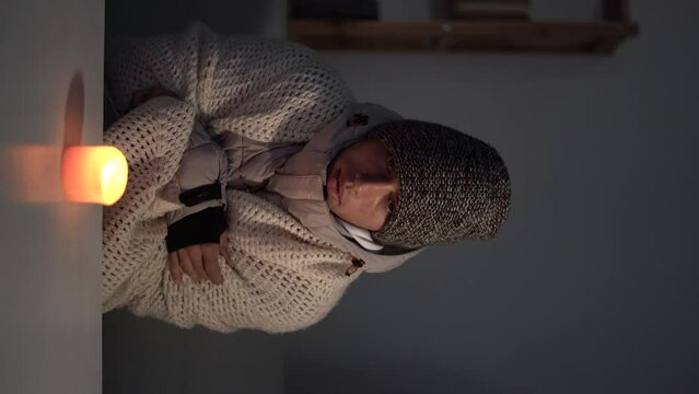 Young frozen man dressed in a warm winter hat and a jacket sits at home at a table and warms his hands from a burning candle. Concept of no heating in winter at home.