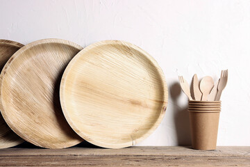 Eco-friendly tableware on wooden table and white wall. Bamboo plates, paper cups and wooden cutlery.