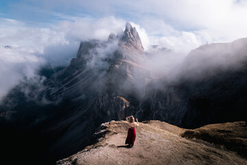 A beautiful girl in a red dress stands facing the mountain partially covered with clouds under the blue sky. Autumn. Italian Alps, Dolomites.