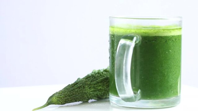 green bitter gourd juice in a transparent glass mug with a whole bitter gourd rotating in white background, side shot