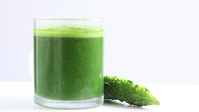 green bitter gourd juice in a transparent glass mug with halved bitter gourd rotating in white background, side shot