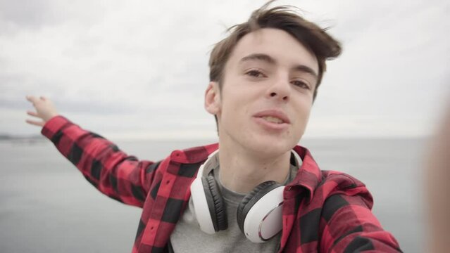 Cute influencer teenager boy talking to camera and holding it on selfie mode. Young man recording travel vlog
