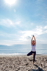 Fototapeta na wymiar Caucasian woman practicing yoga on the seashore looking at the camera. Beautiful fit sporty girl standing full length. Vertical image. Working out concept. Copy space.