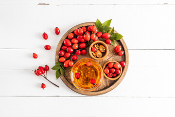 top view of natural rosehip seed oil in a glass bowl on a wooden tray and white wooden table among ripe rose hips. vitamsin C.