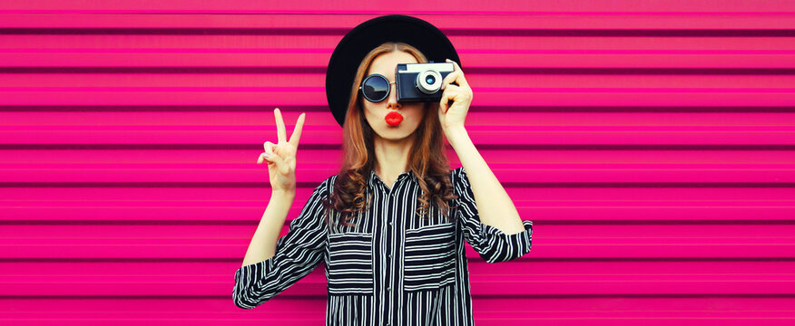 Portrait of stylish modern young woman photographer with film camera blowing her lips on pink background