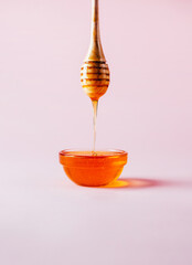 close-up on a wooden spoon with honey dripping into a glass bowl on a pink background