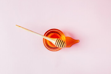 top view of honey in a glass transparent bowl with a wooden spoon of honey spilled on a pink background