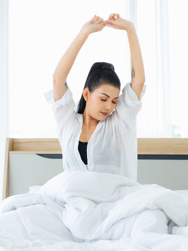 Portrait shot of Millennial Asian sexy female in casual comfort tanktop and shirt sitting under white thick warm blanket posing stretching arm after waking up in bedroom in morning at home.
