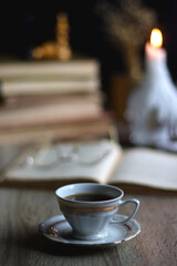 Stack of vintage books, cup of tea or coffee, lit candles, reading glasses and chess pieces on wooden table. Dark academia concept. Selective focus.