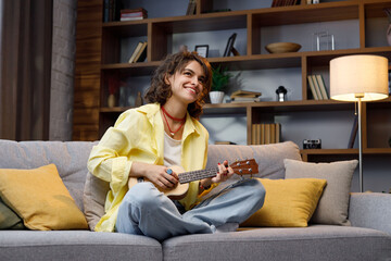 A beautiful hipster girl curly hair in a yellow shirt plays and sings the ukulele on the sofa in the apartment. Home Hobbies playing musical instruments.