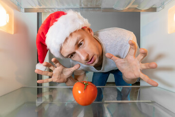 Man in a Santa Claus hat looks suspiciously at a tomato in an empty refrigerator. Concept of an...