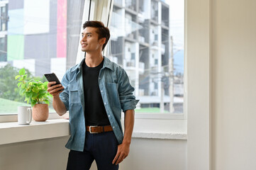 Asian man stands by the window, holding his smartphone and looking through the window.