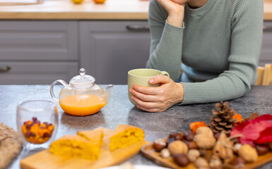 The girl sits alone at the table and drinks a delicious drink from a green mug. The girl is sad in the kitchen alone. On the kitchen table lies a pie and nuts on a wooden board.