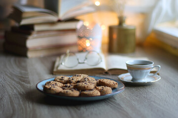 Plate of chocolate chip cookies, stack of vintage books, reading glasses, cup of tea or coffee, lit candle and fairy lights. Hygge at home. Selective focus.