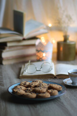 Plate of chocolate chip cookies, stack of vintage books, reading glasses, cup of tea or coffee, lit candle and fairy lights. Hygge at home. Selective focus.