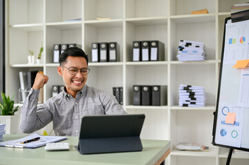 Fototapeta na wymiar Cheerful millennial Asian businessman looking at tablet screen, rejoicing victory, celebrating