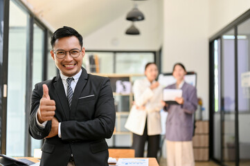 Fototapeta na wymiar Professional adult Asian male boss showing thumb up, standing in his office building